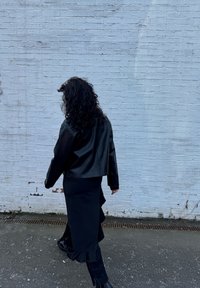 Giacca in pelle nera cropped abbinata a una gonna nera aderente con orlo arricciato. Il modello indossa stivaletti neri contro un muro bianco.