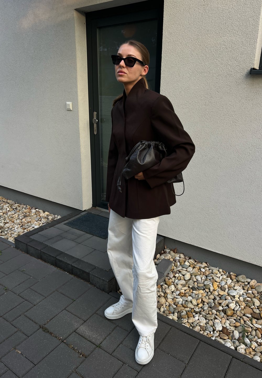 Giacca marrone su misura con colletto alto, pantaloni bianchi larghi e sneakers bianche. Una borsa nera e occhiali da sole completano il look.