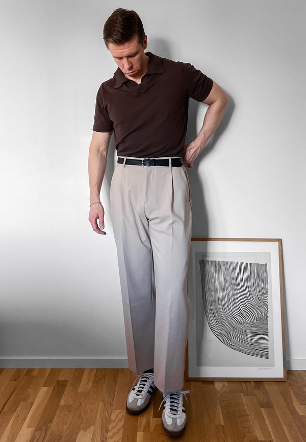 Mand iført mørkebrun polo, beige bukser og sneakers står og kigger ned ved siden af sort-hvidt abstrakt indrammet kunstværk på trægulv.