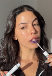 Sinisen metallinhohtoisen huulikiillon levitys kapealla aplikaattorilla, valkoisessa putkessa, jota pitelee henkilö, jolla on pitkät, tummat hiukset ja pisamainen iho.