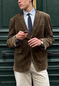 Blazer in velluto a coste marrone con una finitura testurizzata, un bottone singolo e tasche a patta, indossato sopra una camicia azzurro chiaro e una cravatta scura. Pantaloni beige.