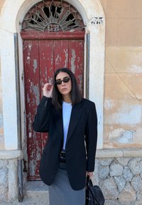 Črn sartorialni blazer preko bele srajce, siva svinčnik krilo in temna sončna očala. Obarvana rdeča lesena vrata služijo kot ozadje.