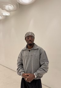 Grauer Pullover mit halbem Reißverschluss, geripptem Kragen und besticktem Logo. Kombiniert mit einer passenden Mütze und goldenen Accessoires. Schlichter Hintergrund.