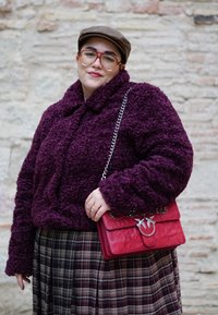 Personne portant une casquette marron, des lunettes rouges, une veste texturée violette, une jupe à carreaux, tenant un sac à main rouge orné d'un oiseau, devant un mur en pierre.