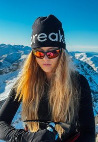 Zwarte muts met de tekst "breaker", spiegelende oranje en rode zonnebril, lang blond haar, zwart shirt met lange mouwen, ijsbijl in de hand, besneeuwde berg op de achtergrond.