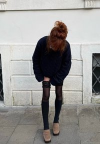 Personne aux cheveux roux en pull noir ajuste des bas en dentelle noire par-dessus des collants noirs, debout sur un pavement en pierre devant un mur blanc avec des fenêtres.