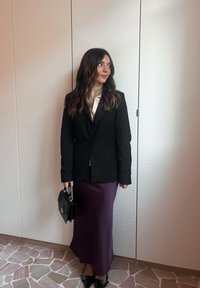 Černé sako přes bílou blůzu, doplněno fialovou midi sukní. Doplňky zahrnují černou kabelku a černé špičaté boty. Hladké textury.