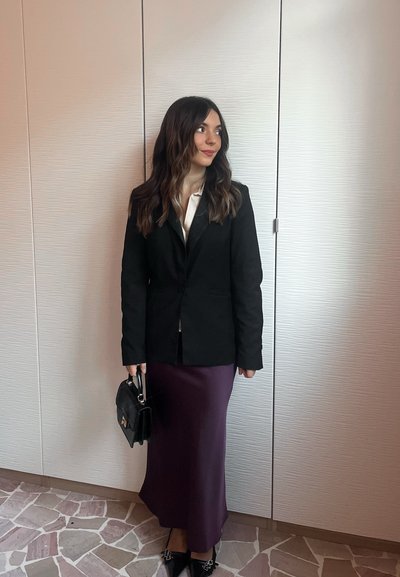 Blazer negra sobre una blusa blanca, combinada con una falda midi morada. Los accesorios incluyen un bolso negro y zapatos negros puntiagudos. Texturas suaves.