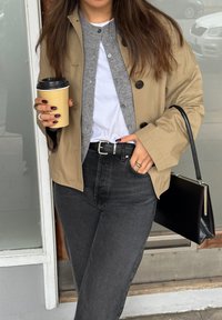Beige jakke over en grå cardigan og hvid skjorte, parret med højtaljede sorte jeans. Holder en engangskaffe-kop og en sort håndtaske.