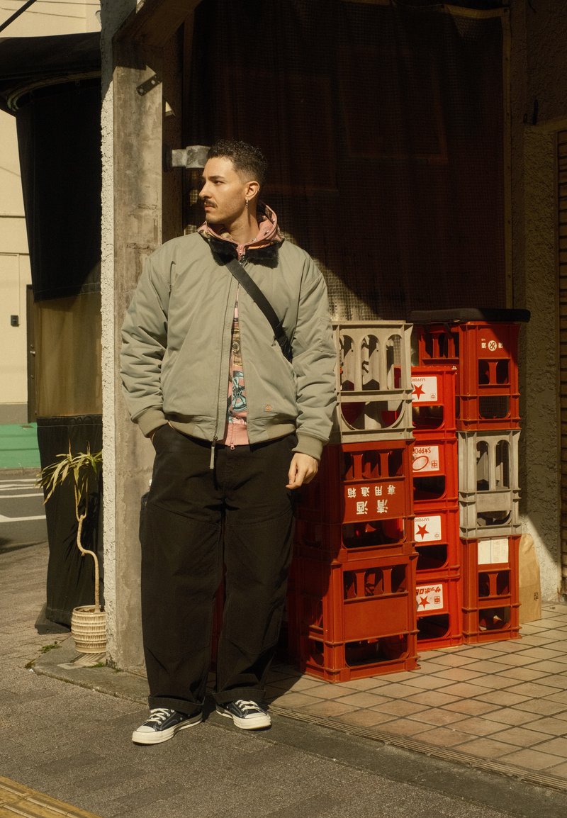 Homme en veste verte et pantalon noir se tient près de caisses en plastique rouges et blanches empilées à l'extérieur d'une entrée de bâtiment ensoleillée.