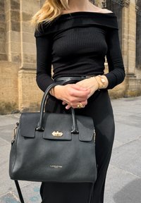 Borsa a tracolla in pelle nera testurizzata con doppi manici, chiusura a scatto e logo dorato. Indossata con un top nero a spalle scoperte a coste e una cintura.