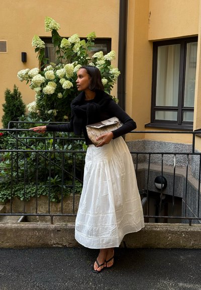 Falda blanca hasta los tobillos con tela texturizada y cintura fruncida. Top ajustado negro, sosteniendo un bolso de mano con estampado. Plantas verdes y fondo de pared beige.