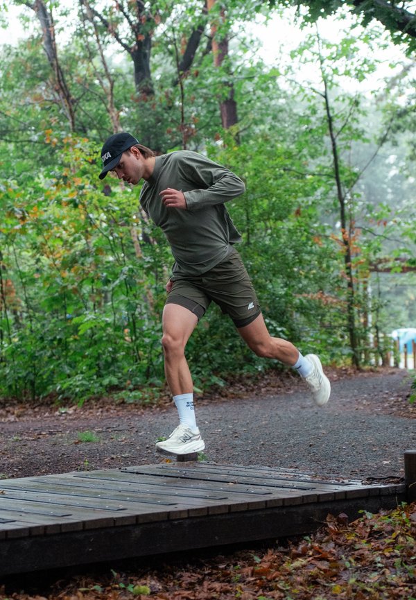 Un hombre vestido para correr salta por encima de un puente de madera en un sendero forestal.