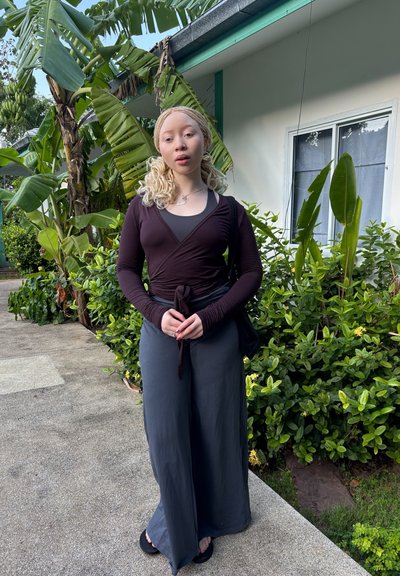 Joven con cabello rubio trenzado que lleva una blusa oscura envolvente y pantalones anchos grises, de pie en un camino junto a plantas tropicales verdes y árboles de plátano.