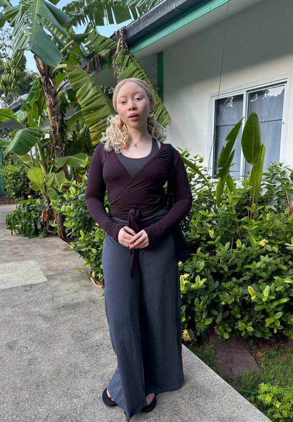 Mladá žena s blonďatými vlasy spletenými do copů, oblečená v tmavém zavinovacím topu a širokých šedých kalhotách, stojí na cestě vedle zelených tropických rostlin a banánovníků.