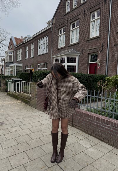Joven mujer con una chaqueta de cuadros marrón y botas, de pie en una acera frente a casas adosadas de ladrillo con vegetación.