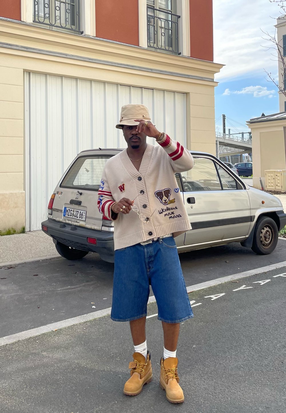 Beige cardiganini punase ja sinise varrukatega, millel on loomade plaastrid. Kanda teksashortside ja kollaste saapade kohal, koos beezist kübara otsaga.