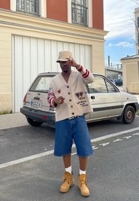 Beige cardiganini punase ja sinise varrukatega, millel on loomade plaastrid. Kanda teksashortside ja kollaste saapade kohal, koos beezist kübara otsaga.