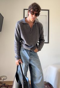 Γκρι πλεκτή μπλούζα με γιακά, σε συνδυασμό με ανοιχτόχρωμα φαρδιά τζιν. Κρατάει μια μαύρη δερμάτινη τσάντα. Μοντέρνα, casual εμφάνιση.