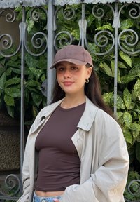 Brun kasket, brun tætsiddende top og lys beige oversized jakke. Baggrunden har grønne blade med en dekorativ metalport.