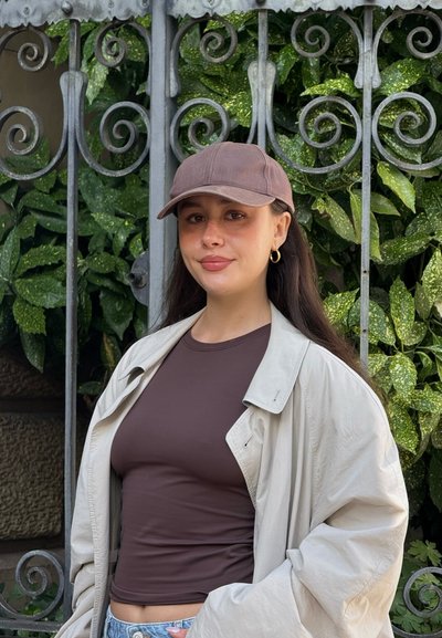 Gorra marrón, top ajustado marrón y chaqueta extragrande beige claro. De fondo, hojas verdes y una puerta de metal decorativa.