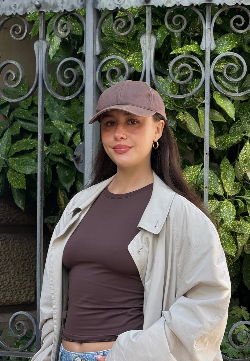 Casquette marron, haut ajusté marron, et veste oversize beige clair. En arrière-plan, des feuilles vertes et une grille métallique décorative.