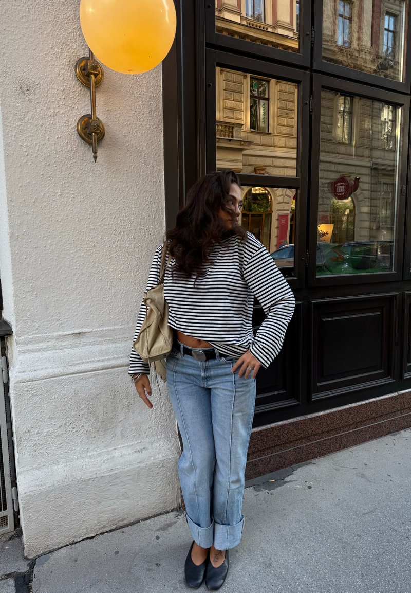 Top a righe bianco e nero con maniche lunghe, jeans azzurri arrotolati, scarpe nere e una borsa beige, con un palloncino giallo sullo sfondo.