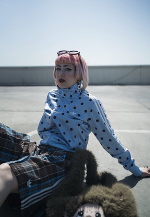 Persona con cabello corto rosa y gafas de sol sentada sobre cemento, vistiendo una chaqueta azul con lunares y pantalones a cuadros, acompañada de un juguete peludo con ojos grandes.