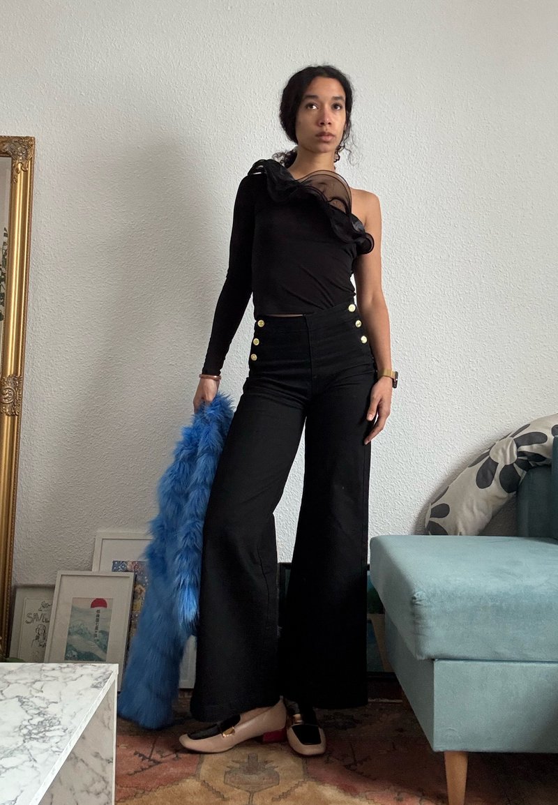 Femme aux cheveux tressés portant un haut noir à une manche et un pantalon noir taille haute, tenant un manteau en fausse fourrure bleu, debout dans le salon.