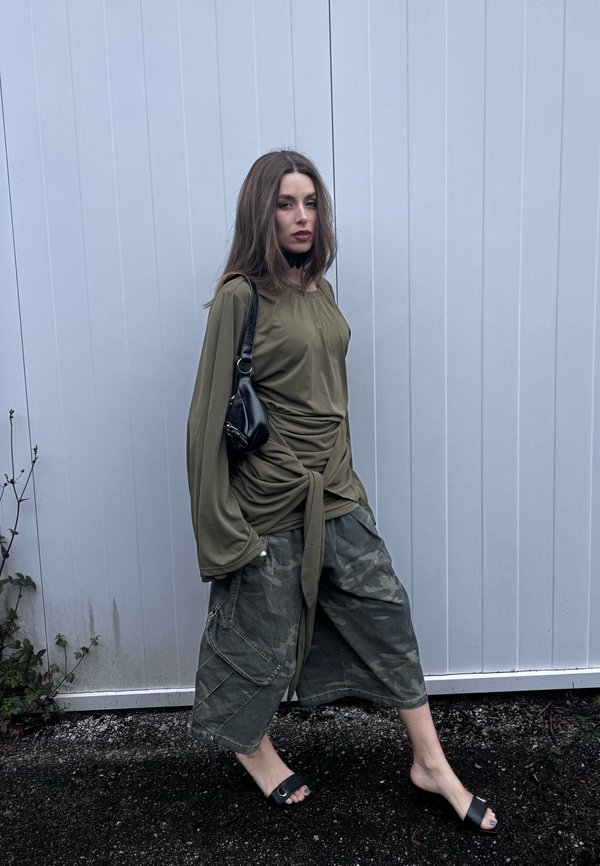 Donna che cammina contro un muro bianco indossando una maglia larga verde oliva, pantaloni larghi mimetici, sandali neri e portando una piccola borsa nera a tracolla.