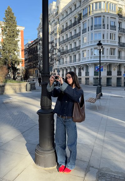 Chaqueta de denim con mangas en contraste, jeans azules, zapatos rojos; sosteniendo una cámara cerca de una farola decorativa en un entorno urbano.