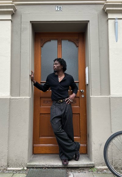 Camisa negra de manga larga, pantalones de pierna ancha a rayas, zapatos negros, de pie junto a una puerta de madera con cristal decorativo, bicicleta cerca.