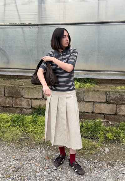 Mujer ajustándose un bolso marrón en el hombro, vistiendo camisa de rayas, falda plisada beige, calcetas rojas y zapatillas negras, de pie sobre grava cerca de un muro de piedra.