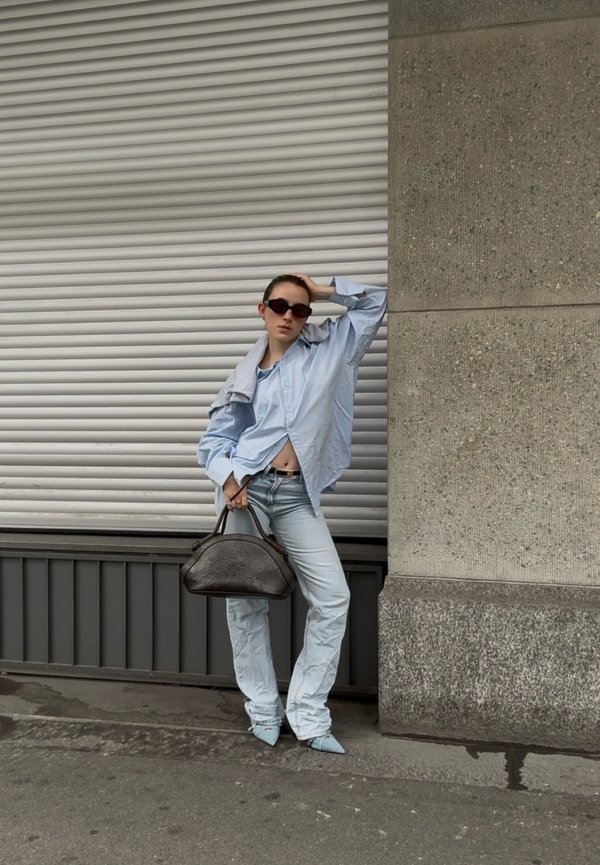Femme portant une chemise oversize bleu clair et un jean, tenant un sac à main foncé, posant devant un volet métallique gris et un mur en béton en extérieur.