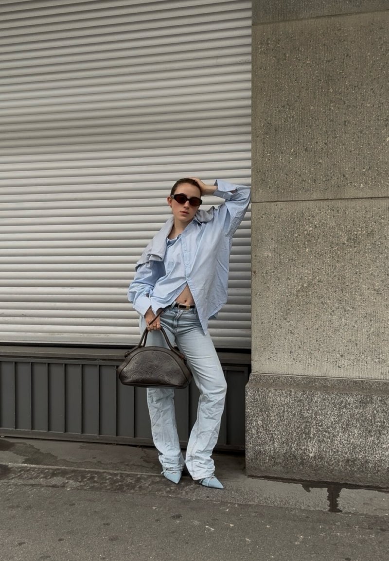 Femme portant une chemise oversize bleu clair et un jean, tenant un sac à main foncé, posant devant un volet métallique gris et un mur en béton en extérieur.