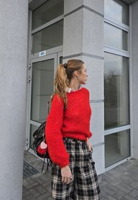 Roter Strickpullover mit strukturiertem Muster, übergroßen Ärmeln, kombiniert mit schwarz-weißen Karohosen. Schwarze Tasche mit pinken Akzenten.