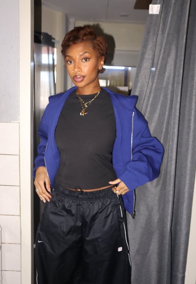 Joven con cabello corto y castaño, vistiendo una sudadera azul con cremallera, top corto negro y pantalones negros holgados, de pie en el interior cerca de una cortina.