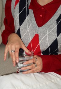 Γκρι και κόκκινο καρό γιλέκο πάνω από κόκκινο πουκάμισο με κολάρο; άτομο holding μια υφασμάτινη γυάλινη με νερό, φορώντας πολλούς ασημένιους δακτυλίους. Λευκό ύφασμα στο παρασκήνιο.
