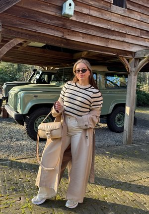 Abrigo trench beige sobre suéter a rayas, pantalones de pierna ancha color crema y zapatillas blancas. Los accesorios incluyen un pequeño bolso beige y gafas de sol.