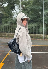 Svijetlosivi korduroy zip-up jakna s kapuljačom, uparena s bijelom majicom i maslinasto zelenim cargo hlačama. Crna torba preko ramena ima detalje s kopčama.