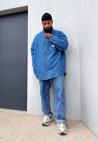 Quilted τζάκετ από τζιν σε μπλε χρώμα, συνδυασμένο με μπλε τζιν και γκρίζα αθλητικά παπούτσια. Σημαντικά χαρακτηριστικά: μεγάλα κουμπιά, ορατή υφή και άνετη εφαρμογή.