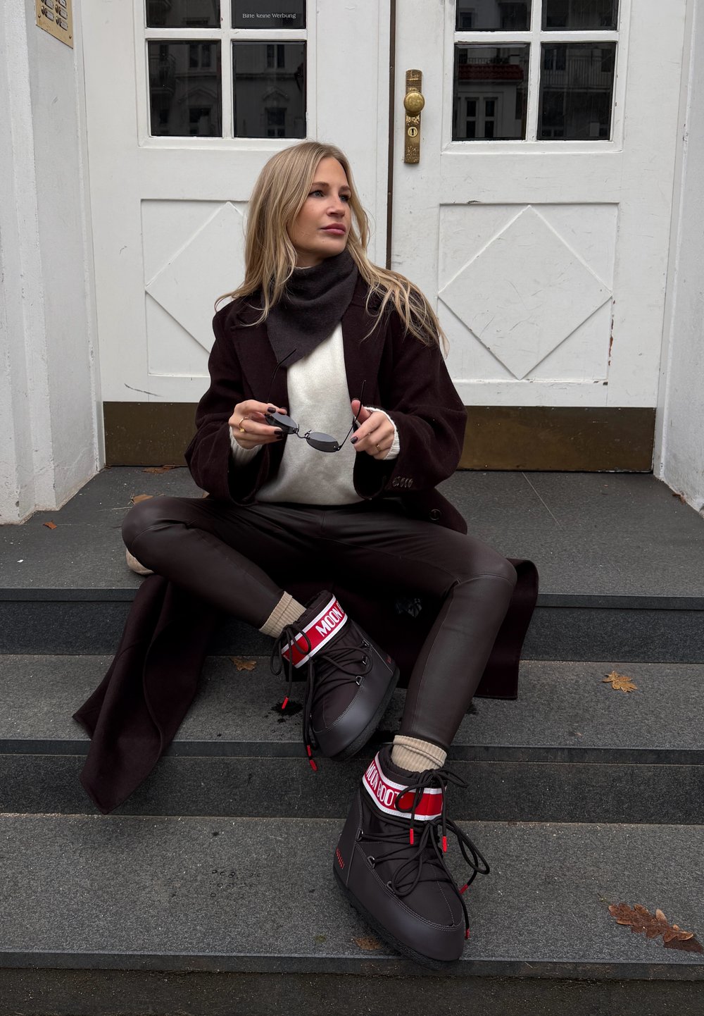 Γυναίκα που κάθεται σε σκαλοπάτια φορώντας καφέ παλτό, δερμάτινο παντελόνι, μπεζ πουλόβερ, κρατώντας γυαλιά  ηλίου, με μαύρες και κόκκινες χειμερινές μπότες με τη λέξη "MOON".