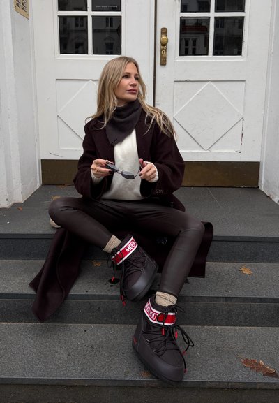 Mujer sentada en unas escaleras con abrigo marrón, pantalones de cuero, suéter beige, sosteniendo gafas de sol y usando botas de invierno negras y rojas con la etiqueta "MOON".
