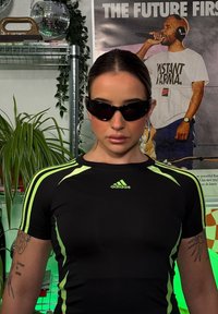 Mustmustreen sportlik särk roheline aktsentidega, tihedalt keha vastav, lühikesed varrukad, koos mustade prillidega. Taustal on taimed ja seina kunst.
