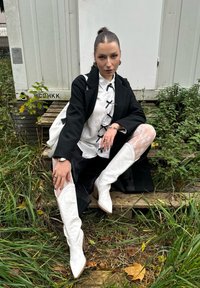 Vita cowboyboots med spetsiga tår och dekorativ sömnad, i kombination med en svart kappa, vit blus och vita spetsstrumpor. Grön växtlighet i bakgrunden.