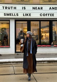 Femme en manteau et béret tenant une tasse de café, debout devant un café avec un panneau sur lequel il est écrit « La vérité est proche et sent le café. »
