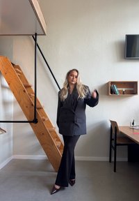 Sacou negru cu două rânduri de nasturi, pantaloni negri evazați și tocuri ascuțite de culoare burgundă. Scară din lemn și decor minimalist în fundal.