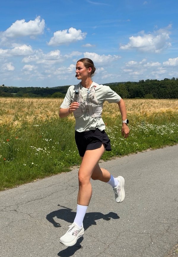 Femme portant une chemise claire et un short noir courant sur un chemin pavé à côté d'un champ sous un ciel partiellement nuageux.