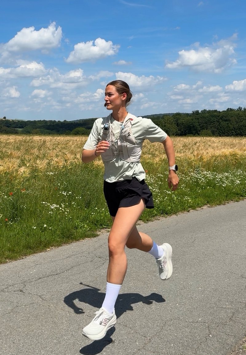 Femme portant une chemise claire et un short noir courant sur un chemin pavé à côté d'un champ sous un ciel partiellement nuageux.