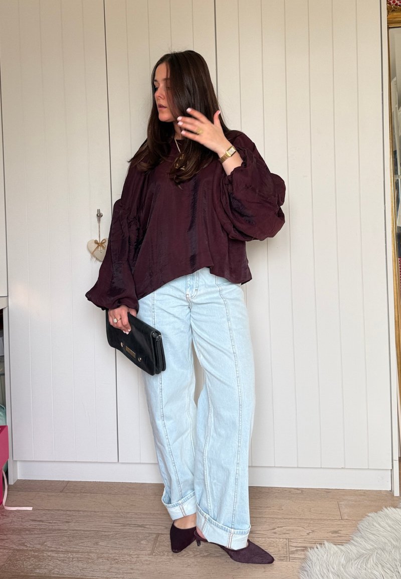 Woman with long brown hair wearing a loose dark purple blouse, light blue wide-leg jeans, purple heels, and holding a black clutch.